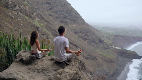 On-a-mountain's-pinnacle,-a-man-and-a-woman-meditate-on-stones,-hands-raised,-while-observing-the-ocean-and-performing-relaxation-breaths